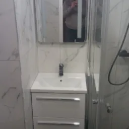 Nowoczesna łazienka z marmurowymi płytkami, białą szafką pod umywalką z dwiema szufladami, lustrem z podświetleniem LED, kabiną prysznicową i czarną marmurową podłogą, widoczne odbicie fotografa...