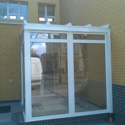 Zewnętrzna zabudowa wejścia do budynku z białych profili aluminiowych i szklanych paneli, z widocznym odbiciem otoczenia i zaparkowanego białego samochodu dostawczego.