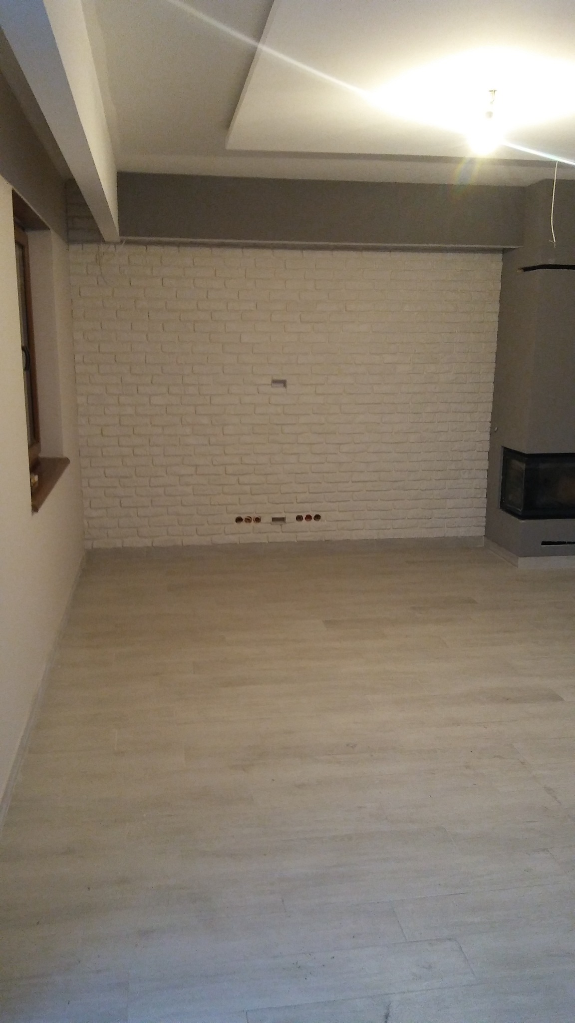 Jasne, przestronne wnętrze z imitacją ceglanej ściany, drewnopodobną podłogą i nowoczesnym kominkiem narożnym.