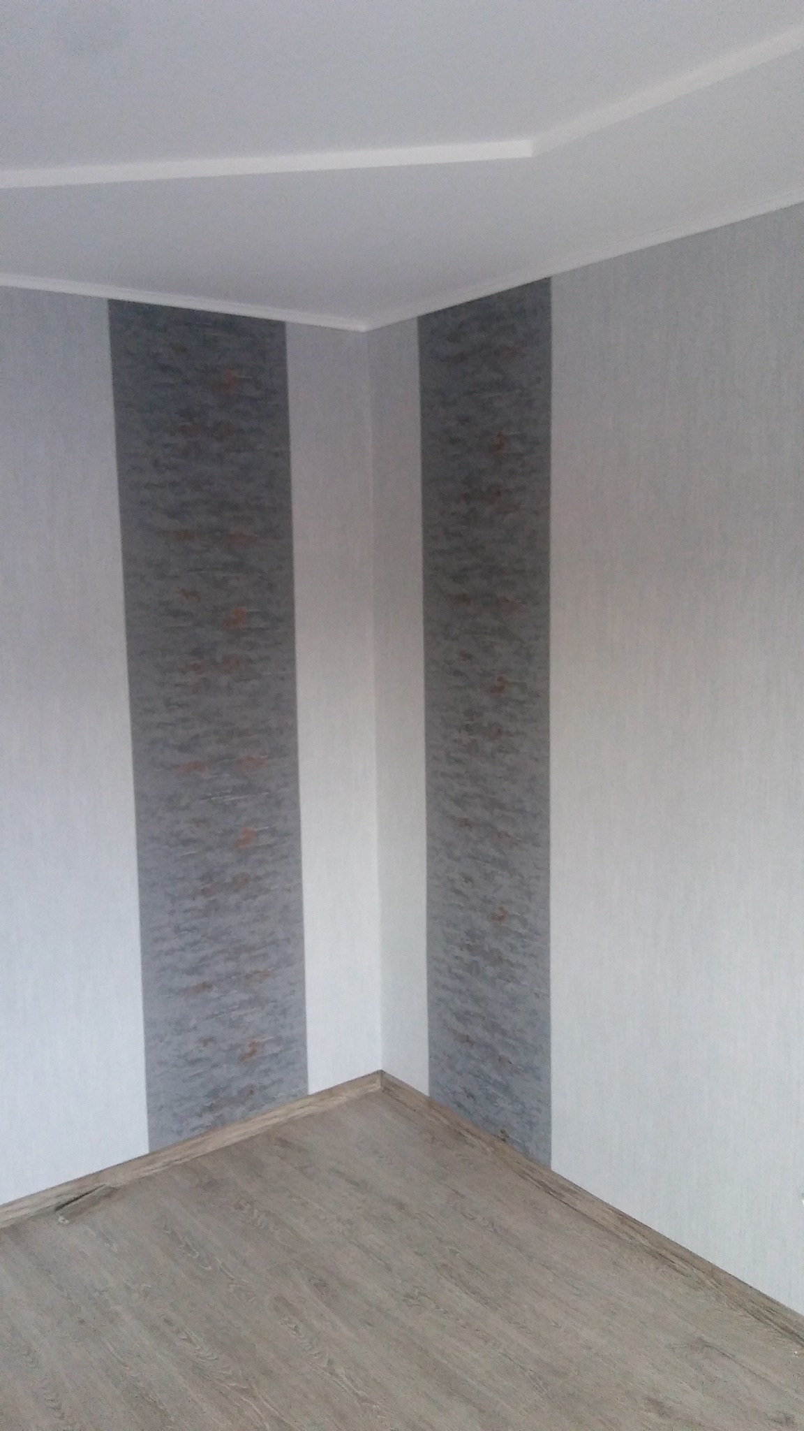 Fragment pokoju po odświeżeniu: pionowe pasy tapety w odcieniach szarości i bieli, jasna podłoga imitująca drewno, widok z dołu.