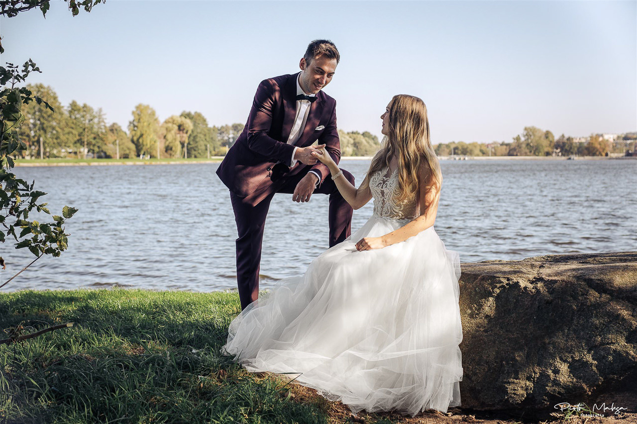 Para młoda nad jeziorem: pan młody w bordowym garniturze opiera się o skałę, trzyma dłoń panny młodej w białej sukni ślubnej z koronkową górą, w tle woda i drzewa.