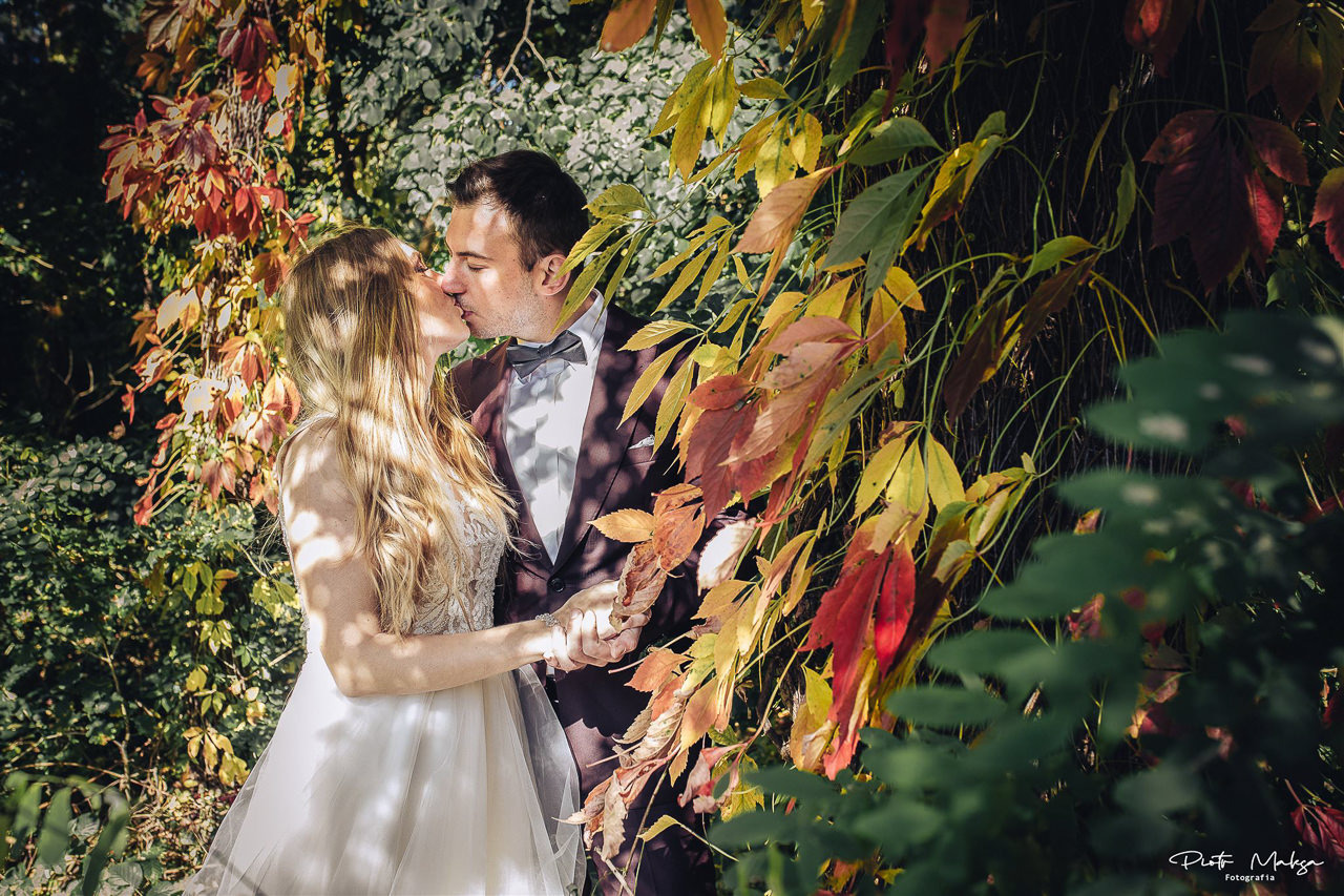 Pocałunek młodej pary w jesiennej scenerii, panna młoda w sukni z koronkową górą, pan młody w bordowym garniturze i muszce, liście w odcieniach czerwieni, żółci i zieleni.