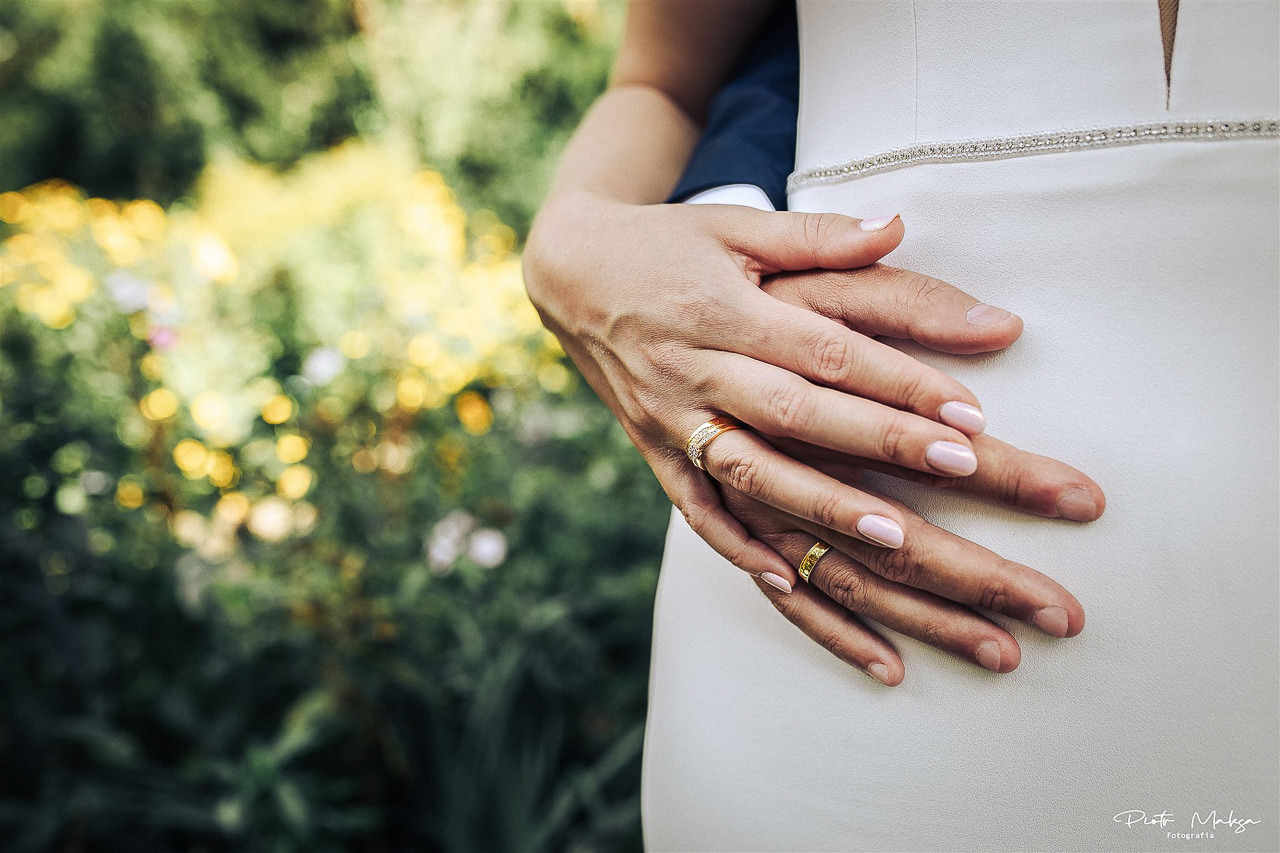 Dłonie pary młodej z obrączkami na tle białej sukni i rozmytego ogrodu.