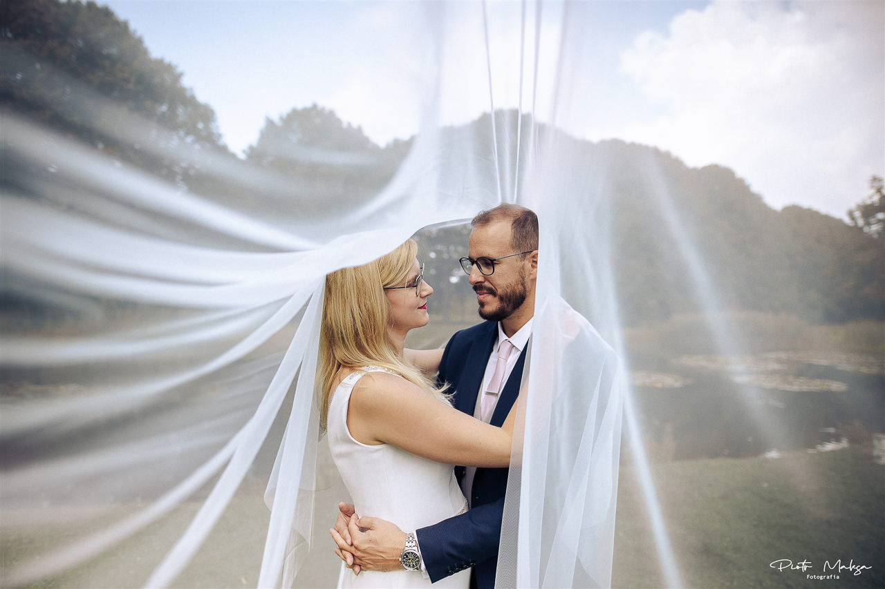 Para młoda otulona białym welonem na tle zieleni parkowej, oboje w okularach, patrzą na siebie z uśmiechem.