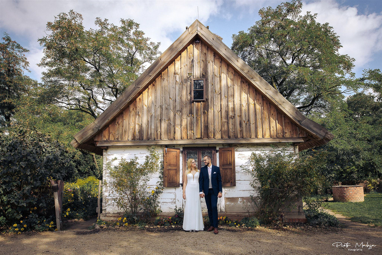Para młoda w eleganckich strojach pozuje przed starą, drewnianą chatą z białymi ścianami i brązowymi okiennicami, otoczoną zielenią drzew i krzewów w plenerowej scenerii.