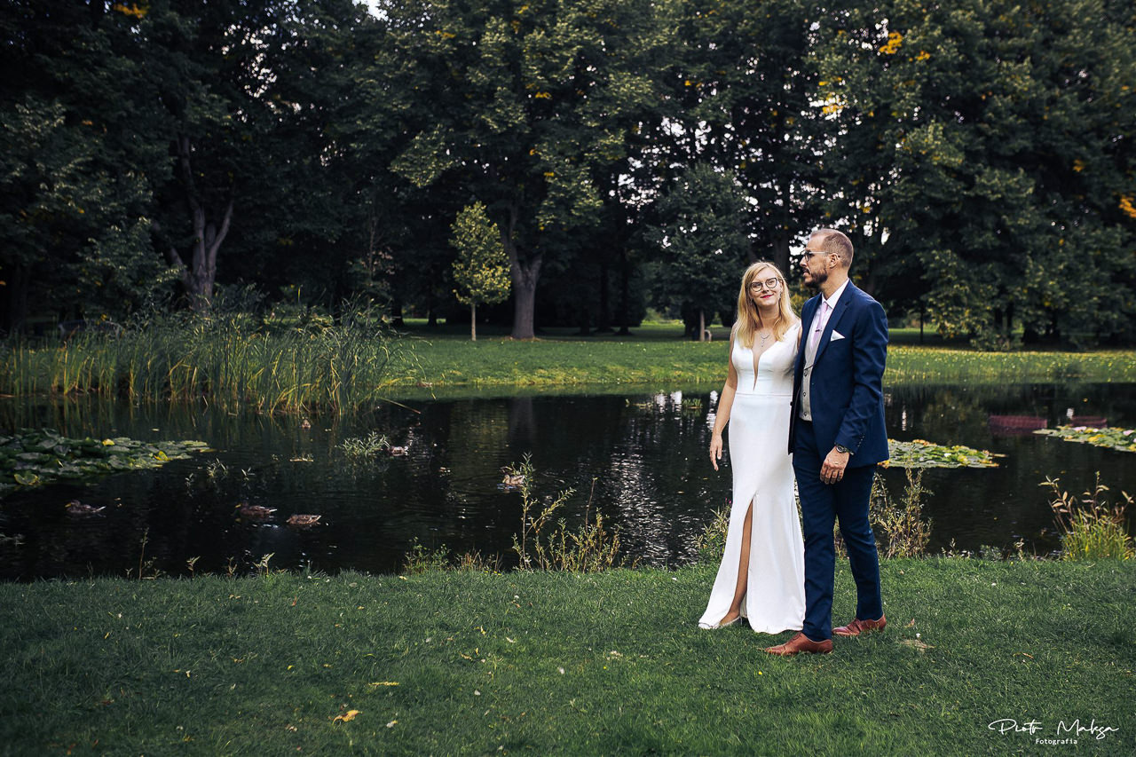 Para młoda w eleganckich strojach pozuje na tle malowniczego stawu z liliami wodnymi i kaczkami, otoczonego bujną zielenią parku.