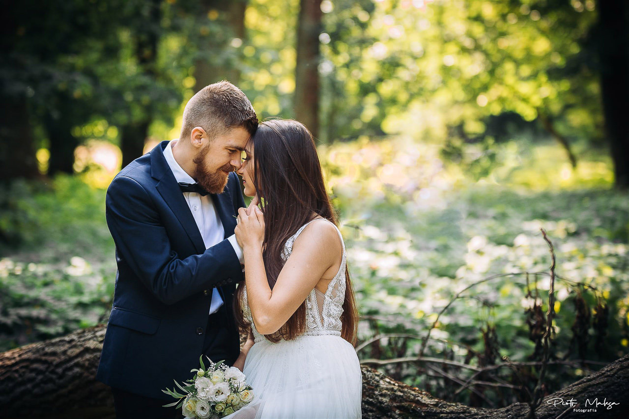 Para młoda w plenerze, delikatny uśmiech, ona w sukni z koronkową górą, on w granatowym garniturze z muszką, bukiet białych róż, leśne tło z prześwitami słońca.