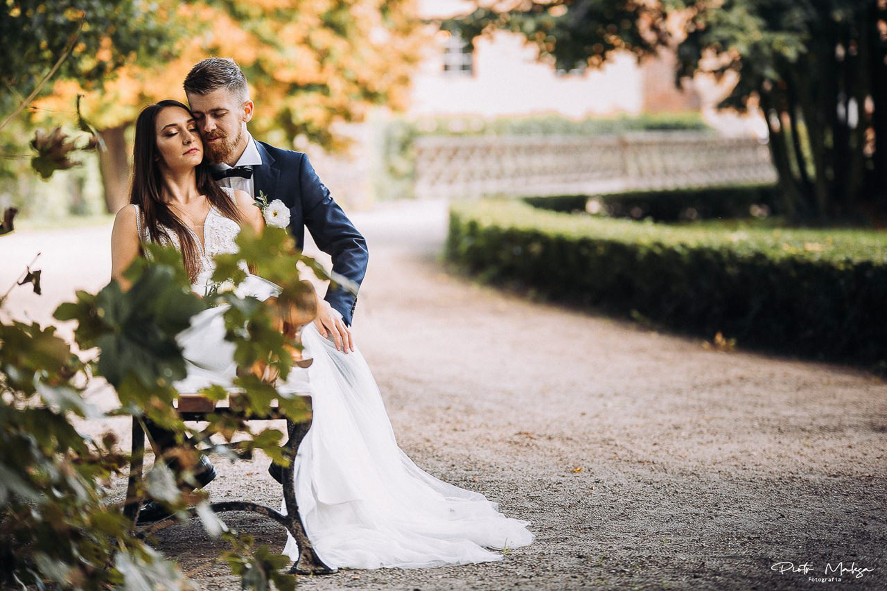 Para młoda siedzi na ławce w parku, panna młoda w białej sukni z koronkową górą, pan młody w granatowym garniturze z muszką, jesienne liście na drzewach w tle.