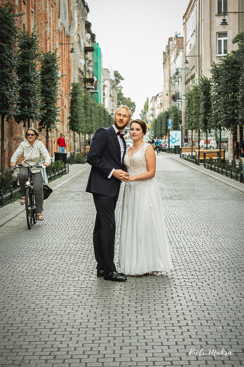 Para młoda w strojach ślubnych pozuje na brukowanej ulicy miejskiej, kobieta na rowerze w tle dodaje dynamiki scenie.