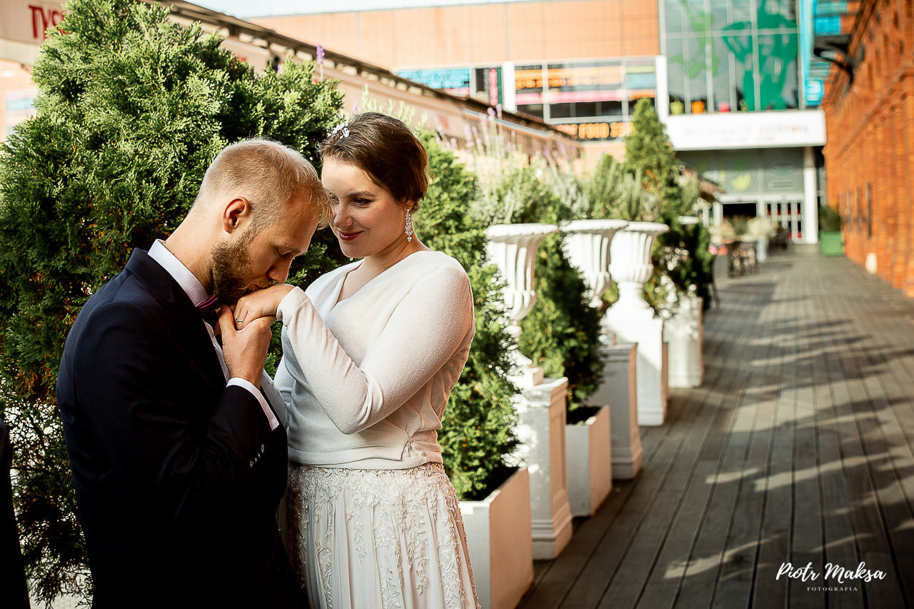 Elegancki mężczyzna w ciemnym garniturze całuje dłoń uśmiechniętej kobiety w koronkowej sukni na tle zielonych krzewów i miejskiej architektury.