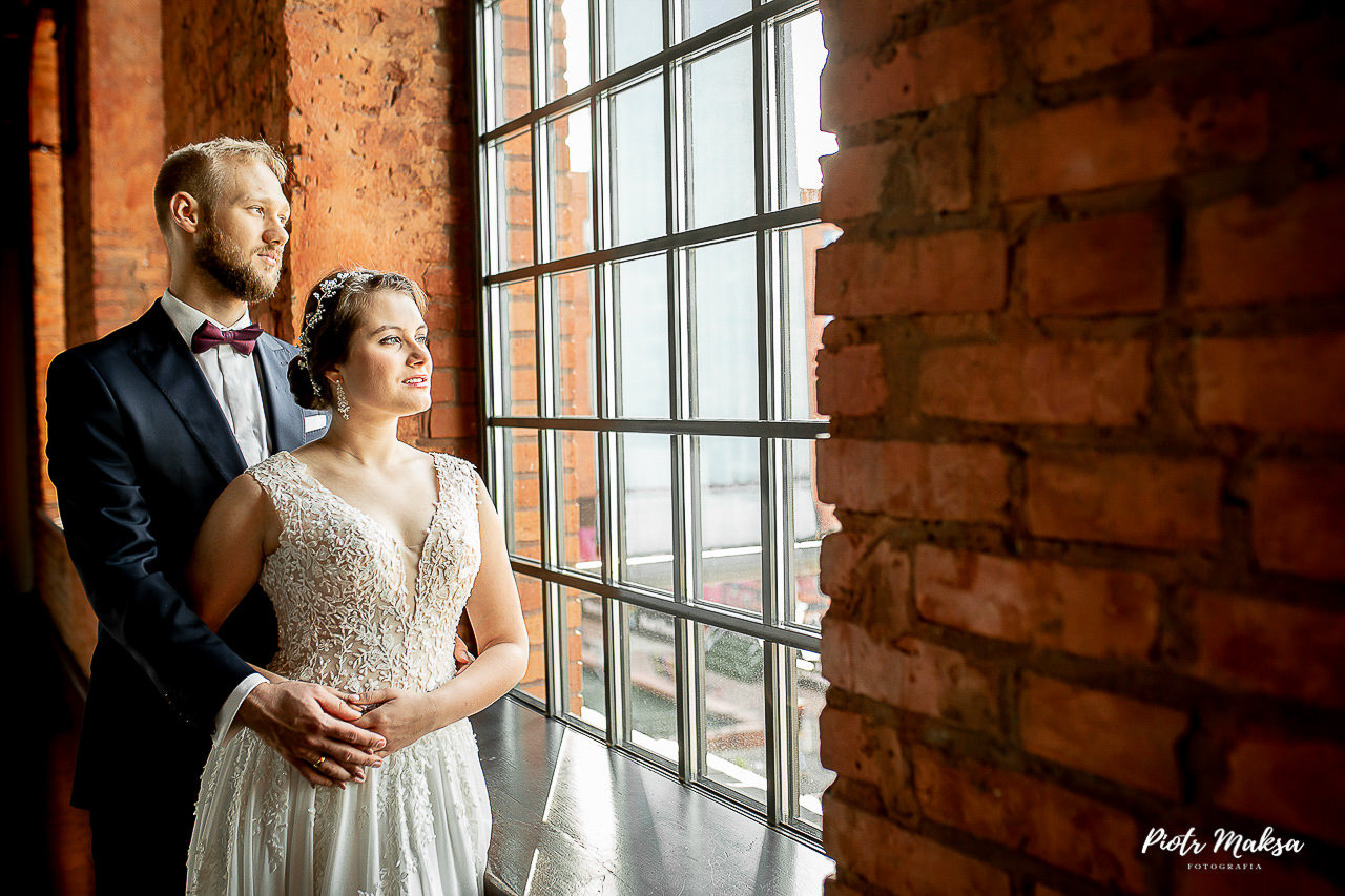 Para młoda w eleganckich strojach pozuje przy oknie w ceglanej scenerii, panna młoda w koronkowej sukni, pan młody w granatowym garniturze z bordową muchą.
