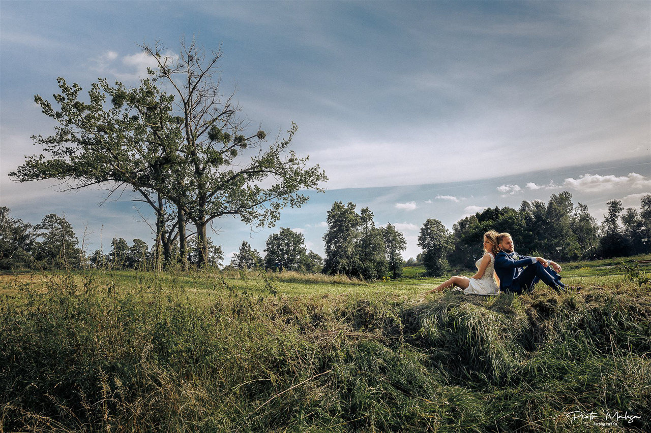 Para młoda siedzi plecami do siebie na skraju łąki z wysoką trawą, w tle samotne drzewo z jemiołą i błękitne niebo z lekkimi chmurami.