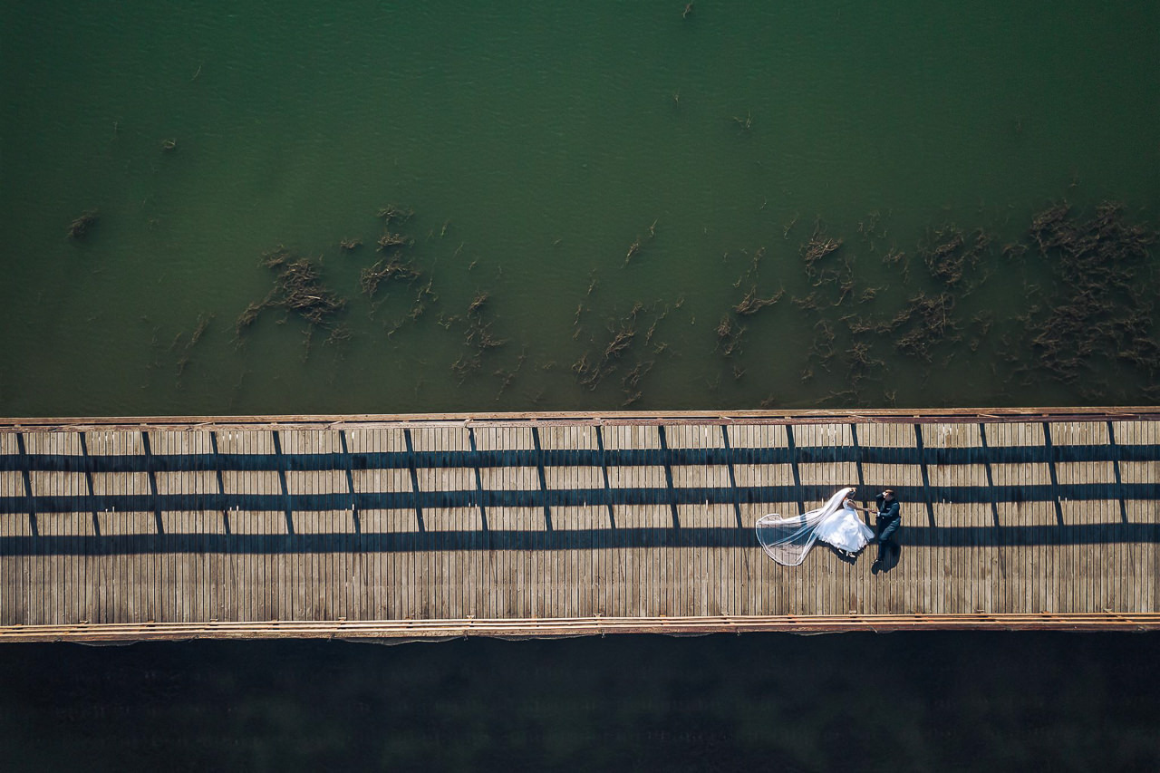 Para młoda leżąca na drewnianym molo, widok z góry, obok ciemna woda z zatopionymi roślinami, cień rzucany przez barierki mola.