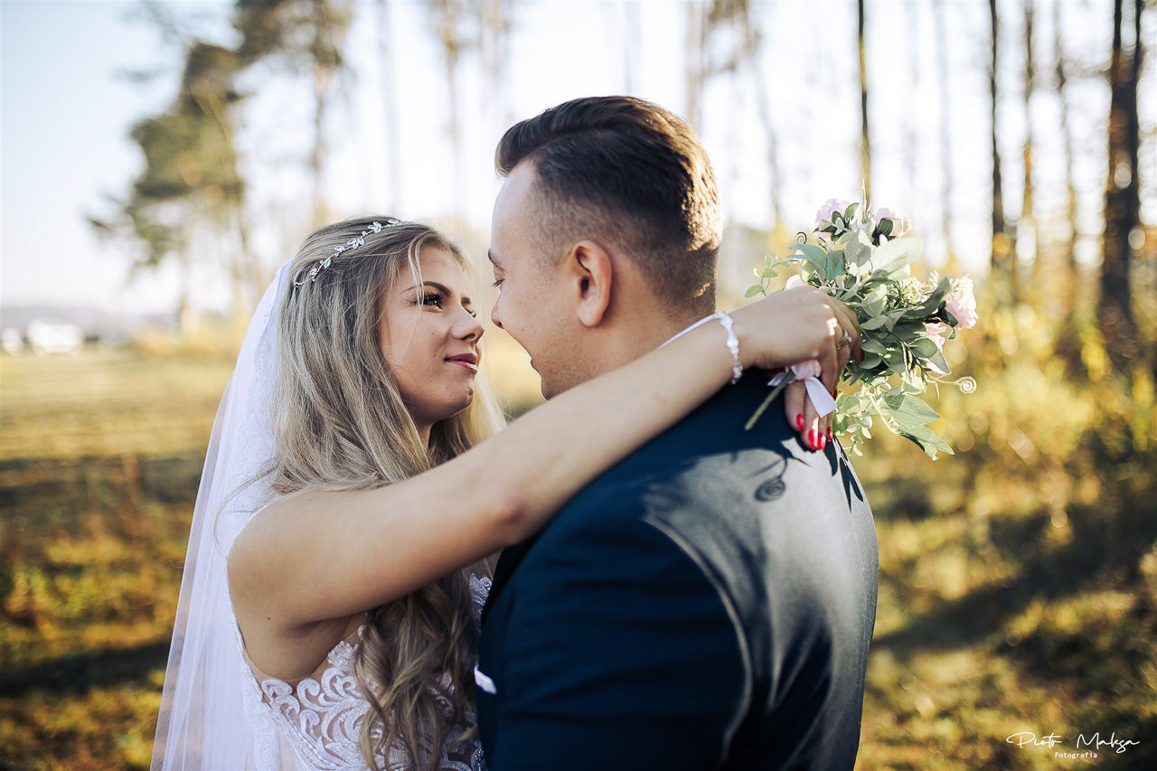 Panna młoda z blond włosami i delikatnym diademem spogląda z uśmiechem na pana młodego, obejmując go za szyję i trzymając bukiet z pastelowych kwiatów.