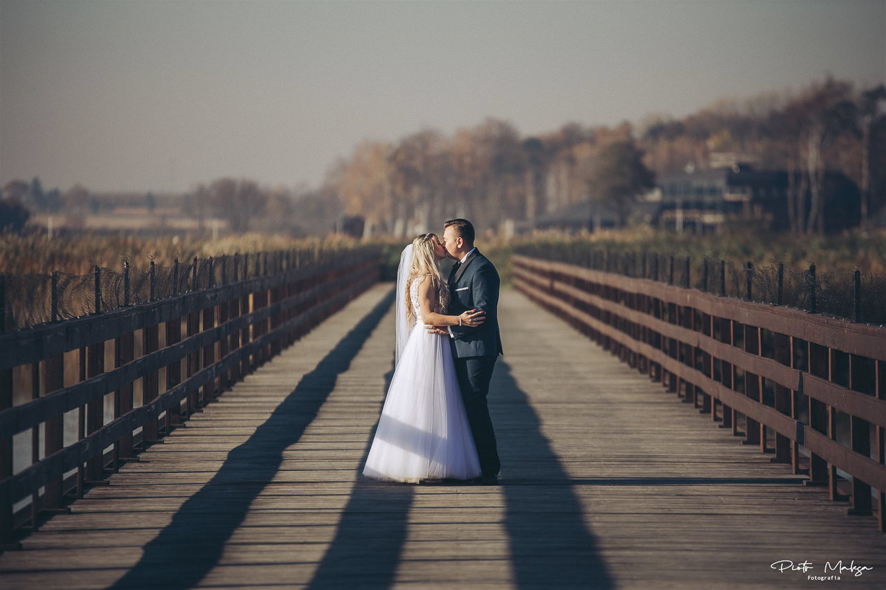 Para młoda całuje się na drewnianym moście z brązową balustradą w słoneczny dzień. W tle widać drzewa i budynek.