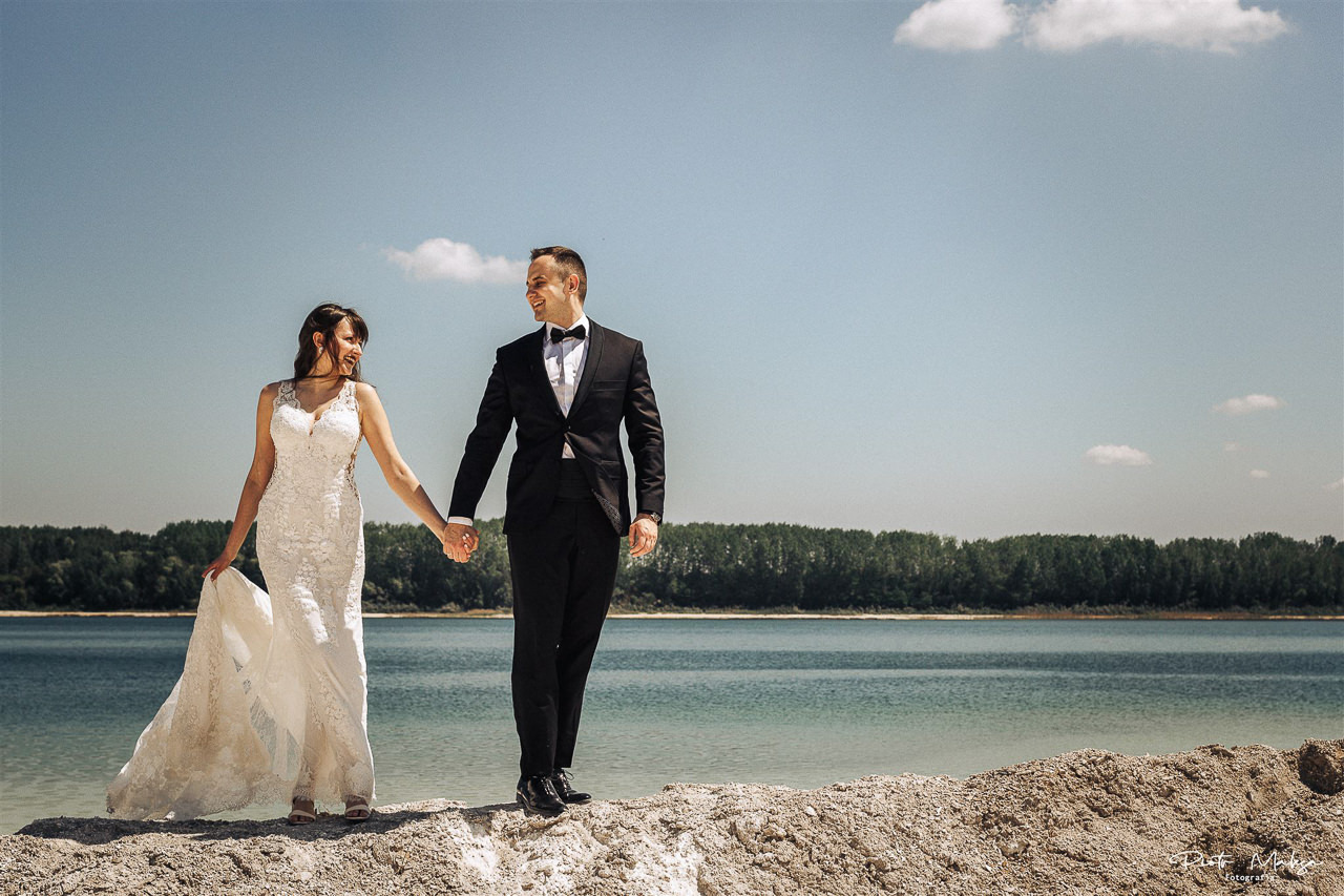 Para młoda, on w czarnym smokingu z muszką, ona w białej koronkowej sukni, trzymają się za ręce na tle błękitnego jeziora i lasu.