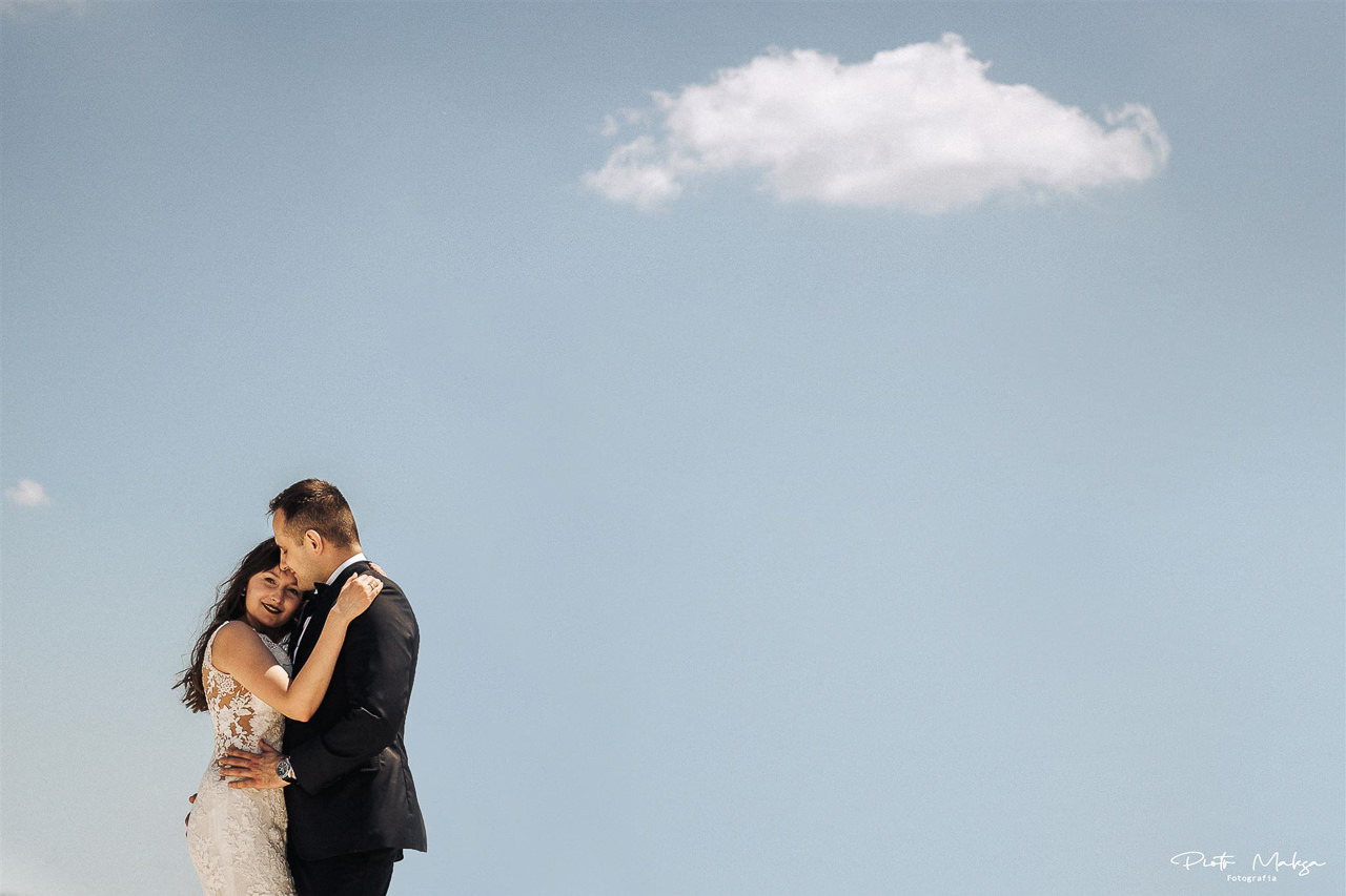 Para młoda w Łodzi przytulona na tle błękitnego nieba z pojedynczą chmurą; panna młoda w koronkowej sukni, pan młody w czarnym smokingu.