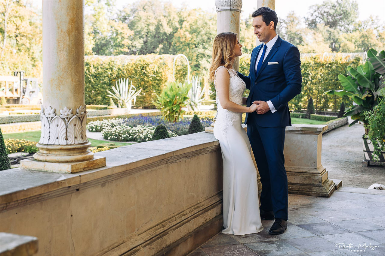 Para młoda trzymająca się za ręce na tle klasycznej architektury i zadbanego ogrodu w słoneczny dzień.