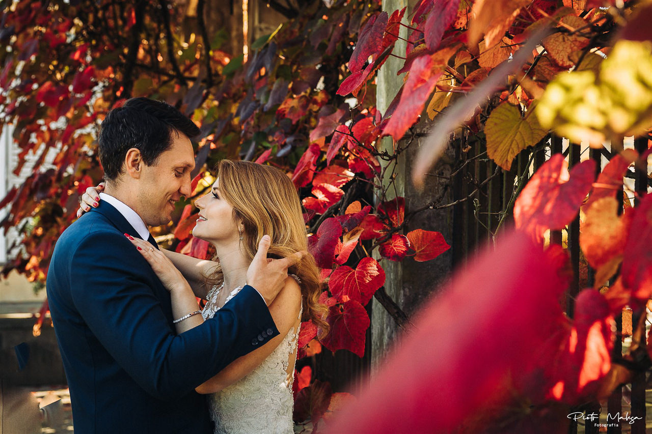 Uśmiechnięta para młoda w objęciach, na tle ściany z czerwonymi liśćmi winobluszczu i fragmentu budynku.