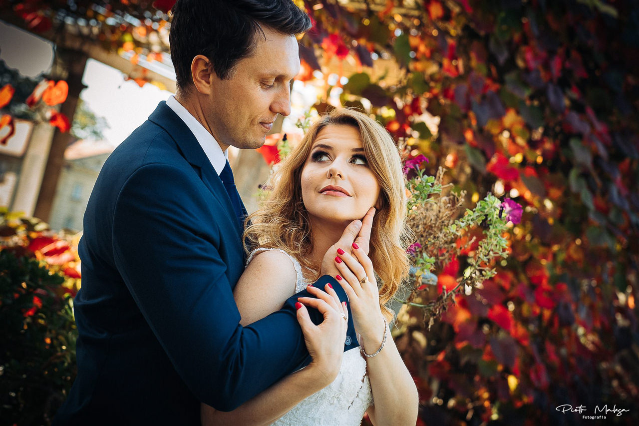 Para młoda w objęciach, panna młoda w białej sukni z koronkową górą, pan młody w granatowym garniturze, tło z kolorowych liści i kwiatów.