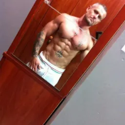 Mężczyzna z tatuażami prezentuje wyrzeźbioną sylwetkę, robiąc selfie w lustrze szafy z drewnianymi drzwiami.