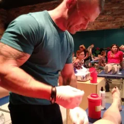 Osoba z tatuażami na ramieniu w rękawiczkach medycznych, pochylona nad pacjentem leżącym na kozetce, w tle inne osoby i kozetki w pomieszczeniu z ceglaną ścianą.