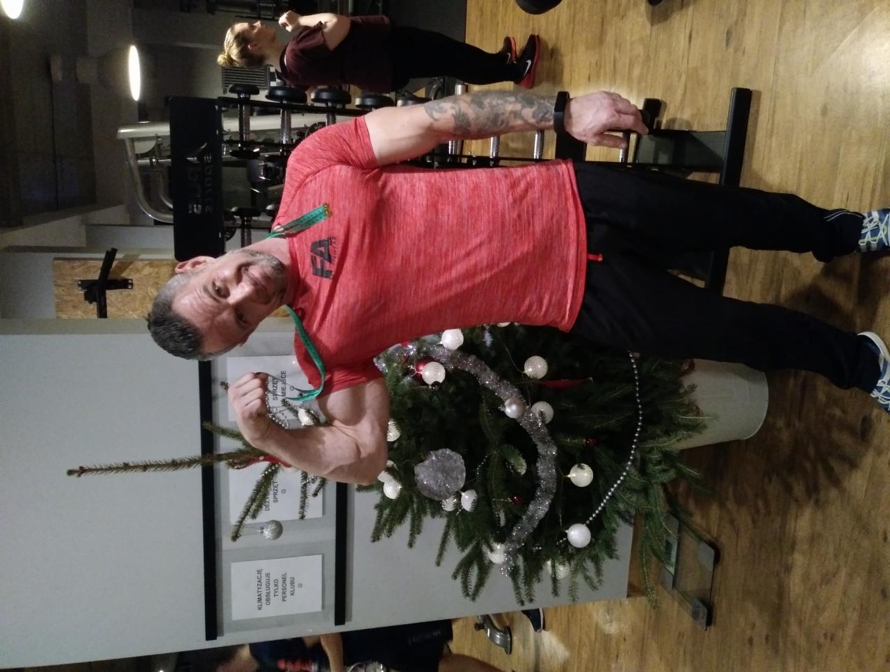 Mężczyzna w czerwonej koszulce prezentuje biceps w siłowni, obok choinki z dekoracjami.
