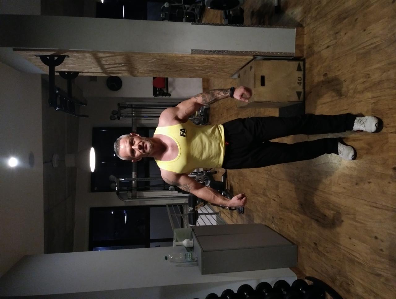 Mężczyzna w żółtej koszulce bez rękawów i czarnych spodniach pozuje na tle siłowni, prezentując muskulaturę i tatuaże na ramionach.