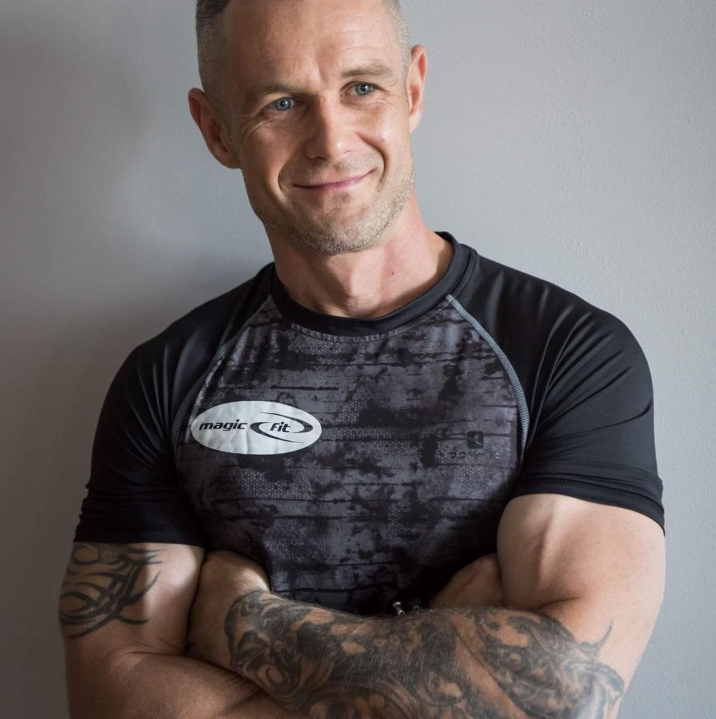 Uśmiechnięty mężczyzna z niebieskimi oczami, w sportowej koszulce z logo 'Magic Fit', z tatuażami na ramionach, stoi na tle szarej ściany z założonymi rękami.