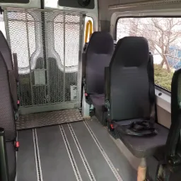 Wnętrze busa przystosowanego do przewozu osób niepełnosprawnych, widoczna platforma wjazdowa i system mocowania wózków inwalidzkich, fotel z pasami bezpieczeństwa.