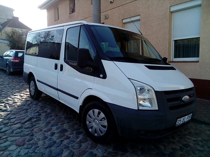 Biały bus Ford Transit na brukowanej ulicy, zaparkowany przed budynkiem z roletami w oknach.