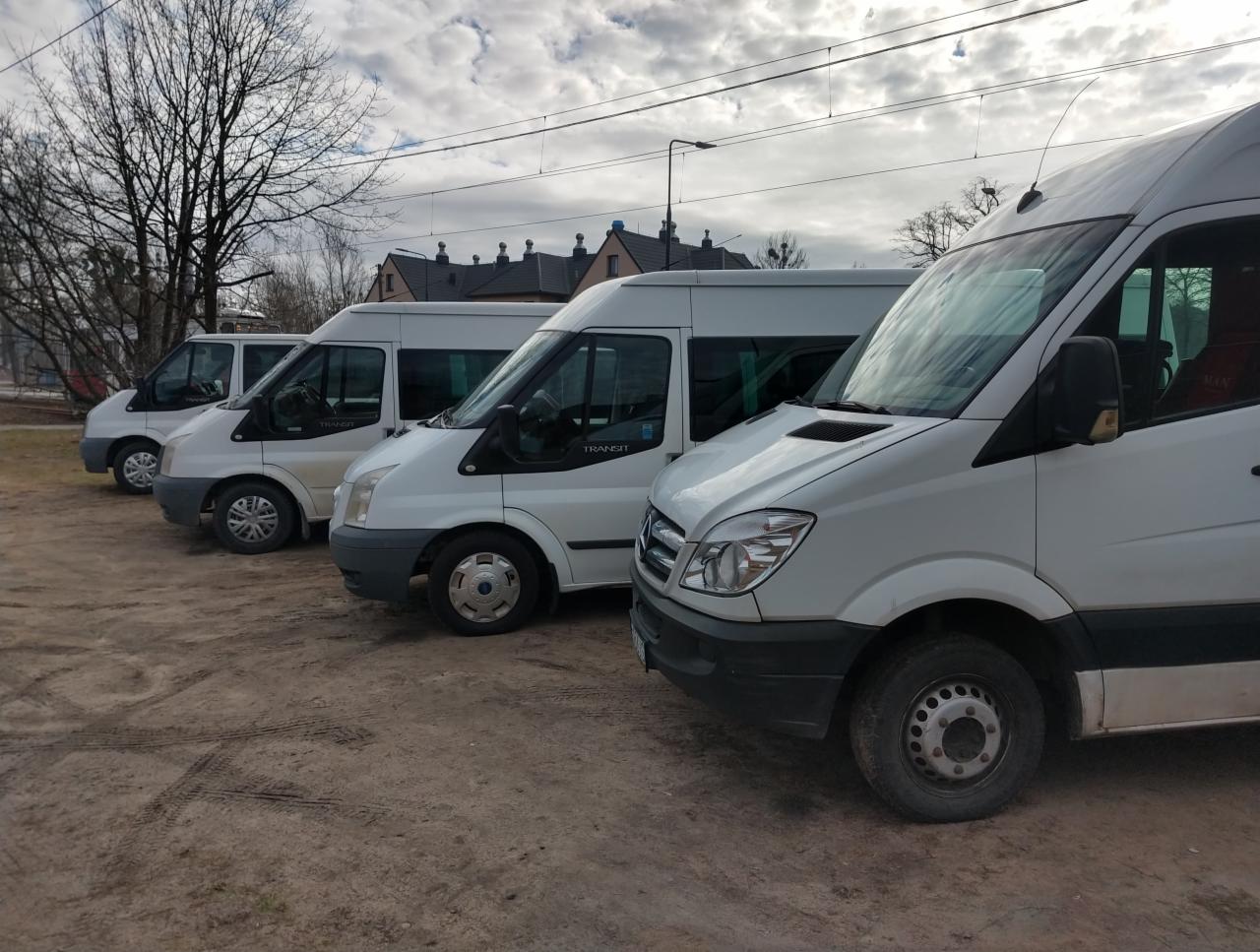 Flota białych busów marki Ford Transit i Mercedes Sprinter zaparkowana na placu z ubitej ziemi, widok z przodu na pojazdy ustawione w rzędzie.