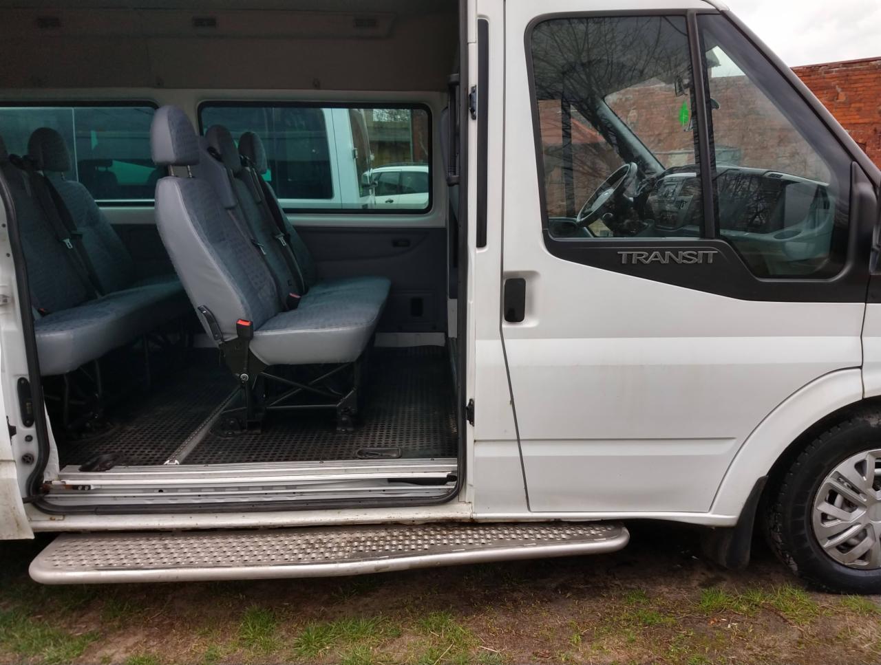 Wnętrze białego busa z otwartymi drzwiami bocznymi, widoczne trzy rzędy szarych siedzeń z pasami bezpieczeństwa, gumowa podłoga i stopień wejściowy.