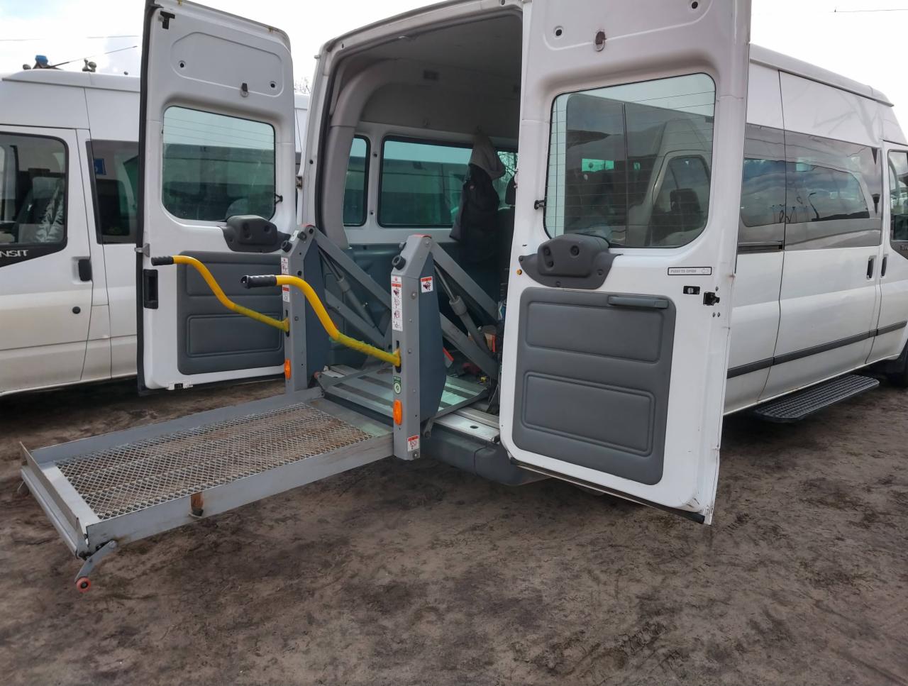 Biały bus z otwartymi drzwiami i rozłożoną platformą dla osób niepełnosprawnych, widoczny mechanizm podnoszący z żółtymi uchwytami.