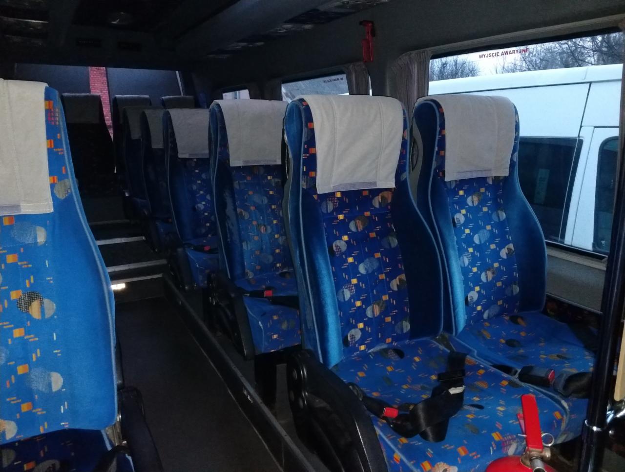 Wnętrze autobusu z rzędami niebieskich foteli z wzorzystą tapicerką i białymi zagłówkami, widoczna gaśnica w dolnym rogu.