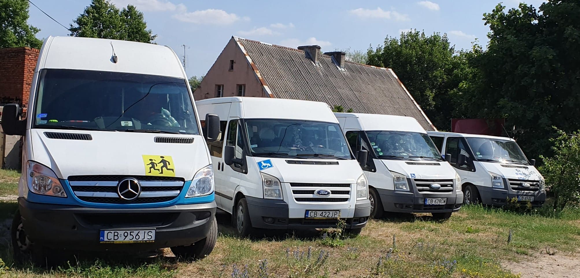 Flota białych busów zaparkowana na trawiastym terenie, pierwszy z niebieskim pasem i symbolem dzieci, drugi z symbolem osoby niepełnosprawnej, w tle budynek z szarą dachówką.