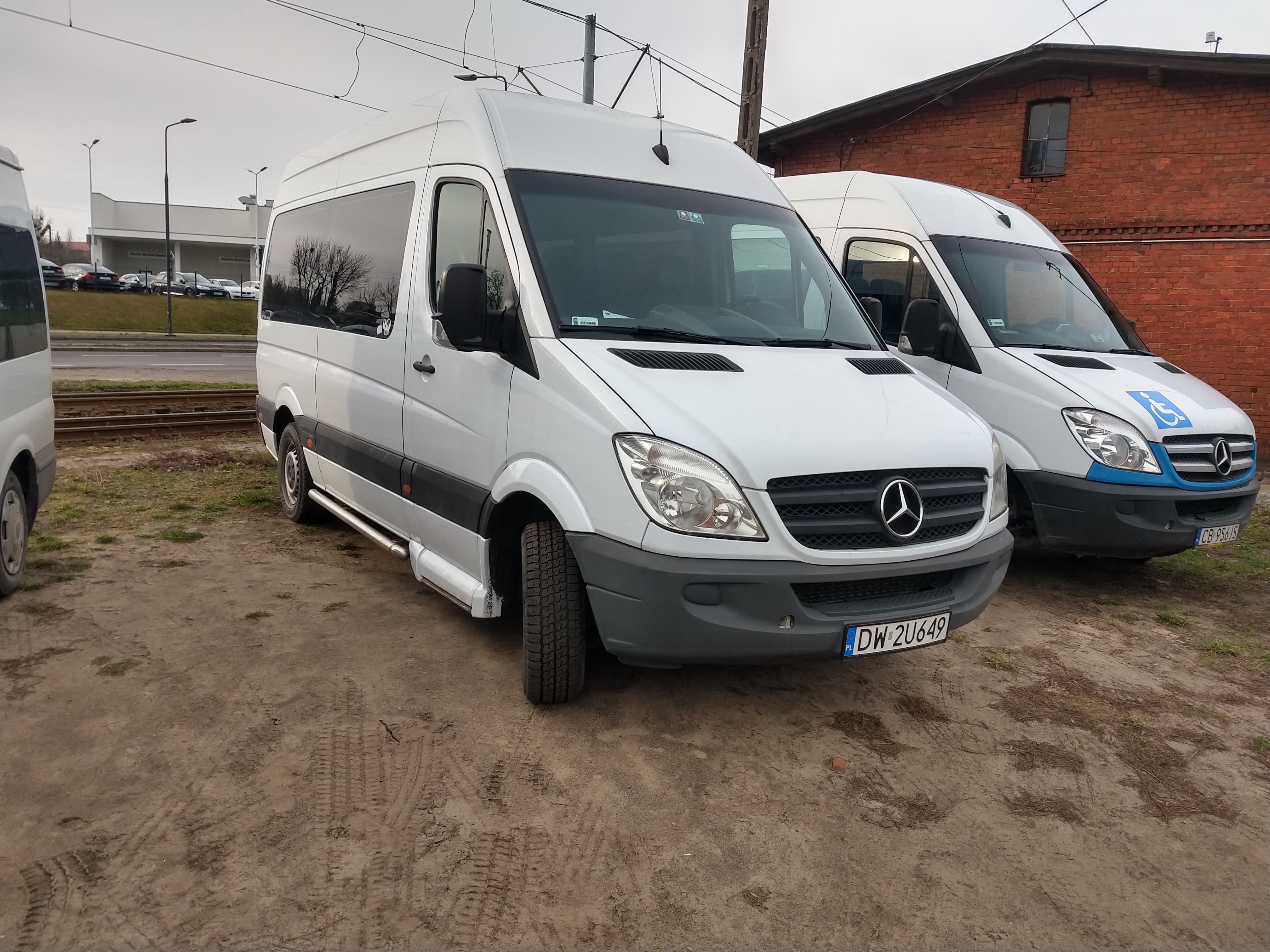 Biały bus Mercedes-Benz z wysuwanym stopniem, zaparkowany na ziemistej powierzchni, z widocznym numerem rejestracyjnym DW 2U649, obok bus z symbolem osoby niepełnosprawnej.