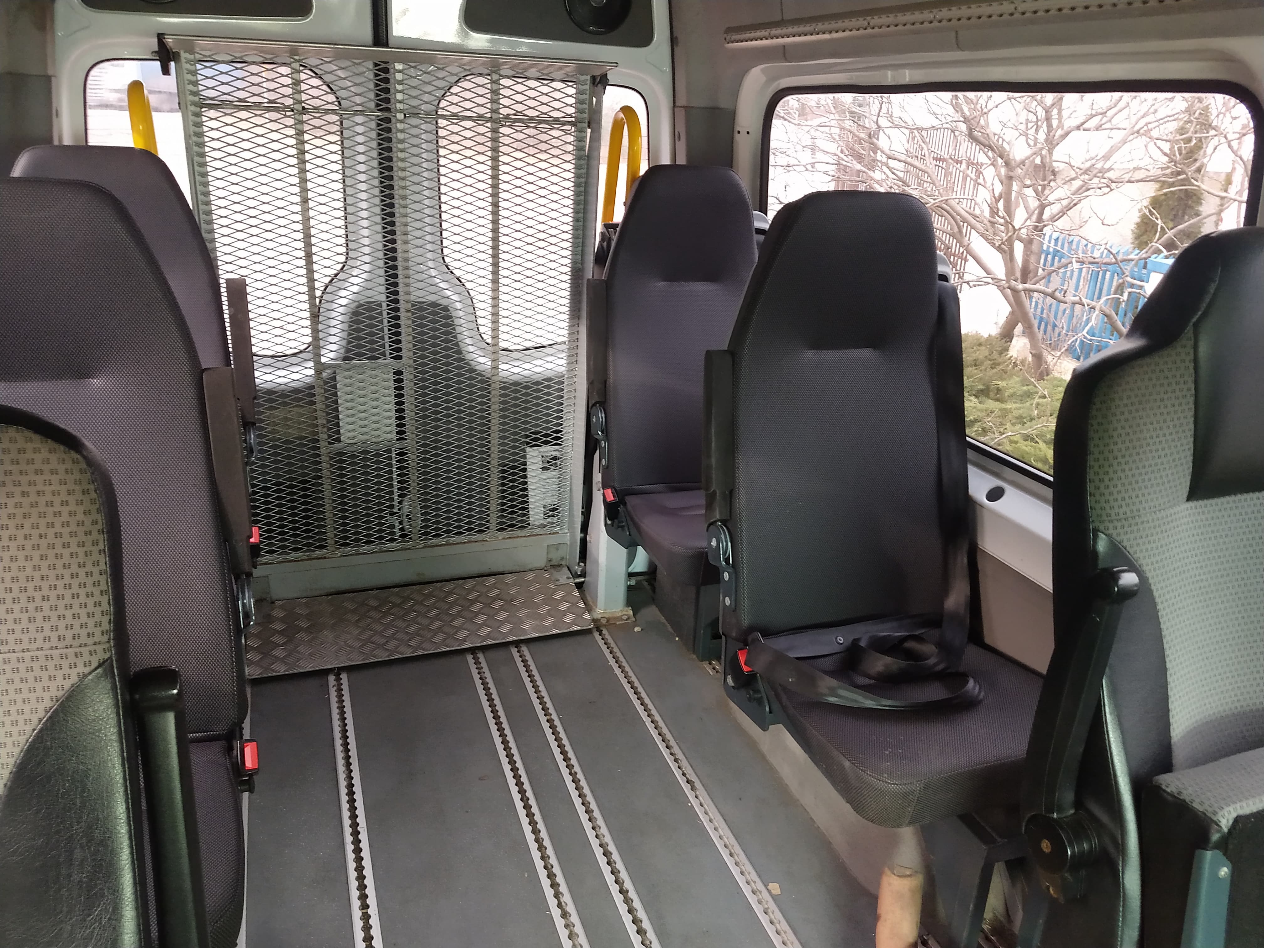 Wnętrze busa przystosowanego do przewozu osób niepełnosprawnych, widoczna platforma wjazdowa i system mocowania wózków inwalidzkich, fotel z pasami bezpieczeństwa.