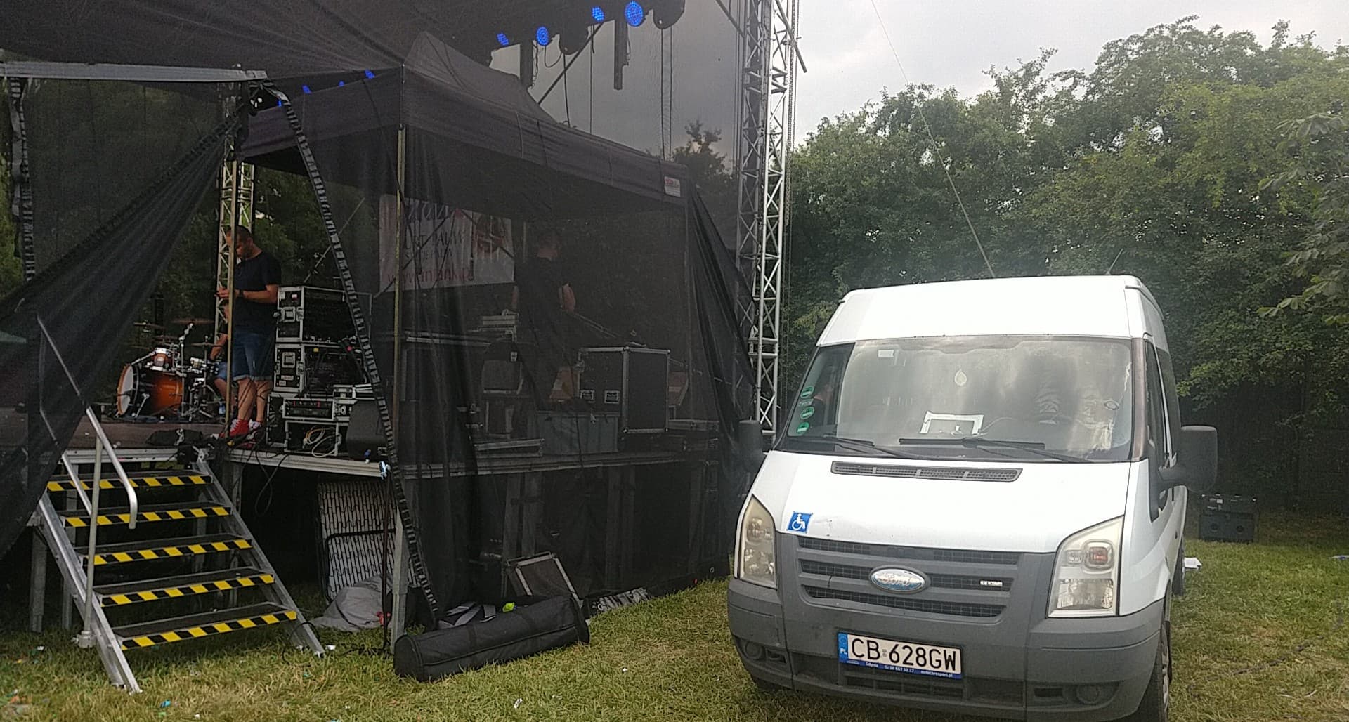 Biały bus z symbolem niepełnosprawności zaparkowany obok sceny z perkusją i sprzętem nagłośnieniowym, przygotowania do koncertu plenerowego.