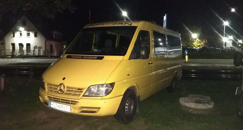 Żółty bus Mercedes-Benz Sprinter z napisem 'DOM-JAN' nad przednią szybą, sfotografowany nocą przy torach kolejowych i budynku.