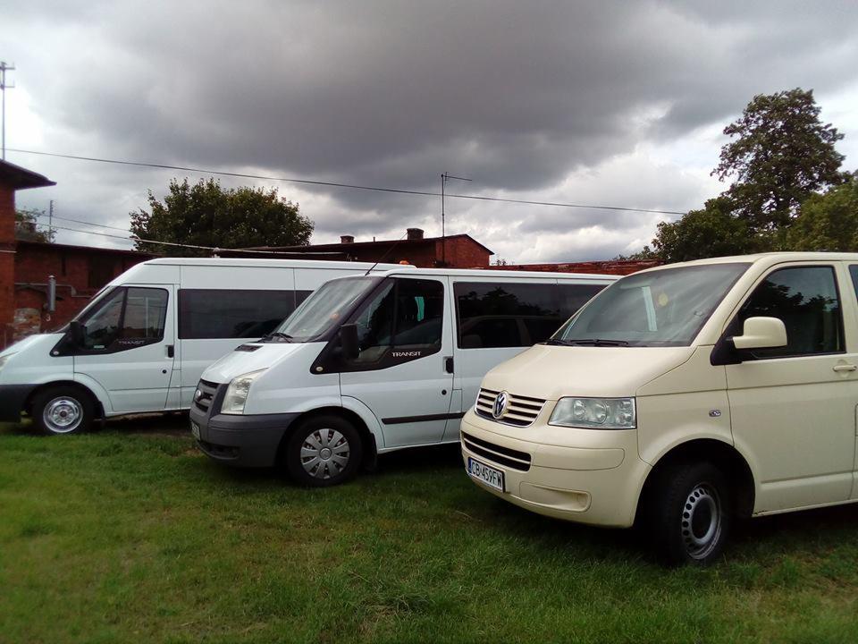 Trzy busy (Ford Transit i Volkswagen Transporter) zaparkowane na trawie przed ceglanym budynkiem pod pochmurnym niebem.