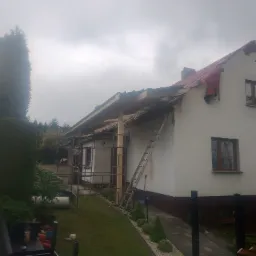 Dom jednorodzinny z częściowo zdjętym poszyciem dachu, widoczne krokwie, zabezpieczony czerwoną folią. Przystawiona drabina i rusztowanie. Pochmurne niebo w tle.