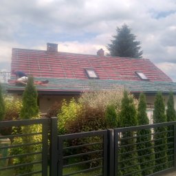 MarBud - Pracownik w trakcie układania czerwonej dachówki na konstrukcji z łat i kontrłat, widoczne okna dachowe i komin, dom otoczony zielenią, ujęcie zza ogrodzenia.