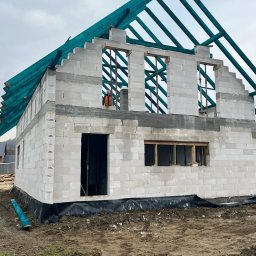 MarBud - Surowy stan domu z betonu komórkowego w trakcie budowy. Widoczne ściany, otwory okienne z drewnianymi ramami, zielona konstrukcja dachu i pracownik w pomarańczowej kamizelce na górze.