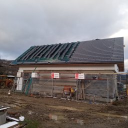 MarBud - Budowa domu murowanego w trakcie układania dachówki; część dachu pokryta, na drugiej części widoczne membrany i łaty; na pierwszym planie rusztowania i materiały budowlane, teren wokół placu budowy...