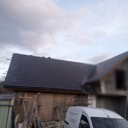 MarBud - Dom w trakcie budowy z częściowo ukończonym, ciemnym dachem z dachówki ceramicznej, widoczny z boku, na pierwszym planie biały samochód dostawczy z danymi firmy.