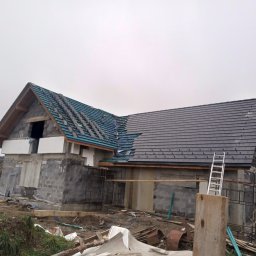 MarBud - Węgierska Górka: Budowa domu z częściowo pokrytym dachem ciemną dachówką, widoczny ruszt pod drugą część dachu i drabina oparta o ścianę budynku.