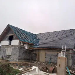 Węgierska Górka: Budowa domu z częściowo pokrytym dachem ciemną dachówką, widoczny ruszt pod drugą część dachu i drabina oparta o ścianę budynku.