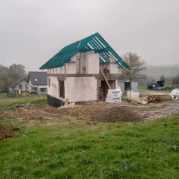 Dom murowany w trakcie budowy, widoczne ściany z bloczków, konstrukcja dachu z zielonymi łatami, materiały budowlane Solbet, teren błotnisty po deszczu.