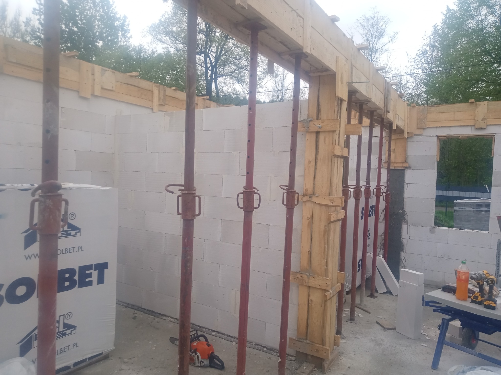 Wznoszenie ścian z białych bloczków, drewniane szalunki podtrzymywane przez stalowe stemple, piła łańcuchowa na podłożu, widoczny materiał budowlany marki Solbet.